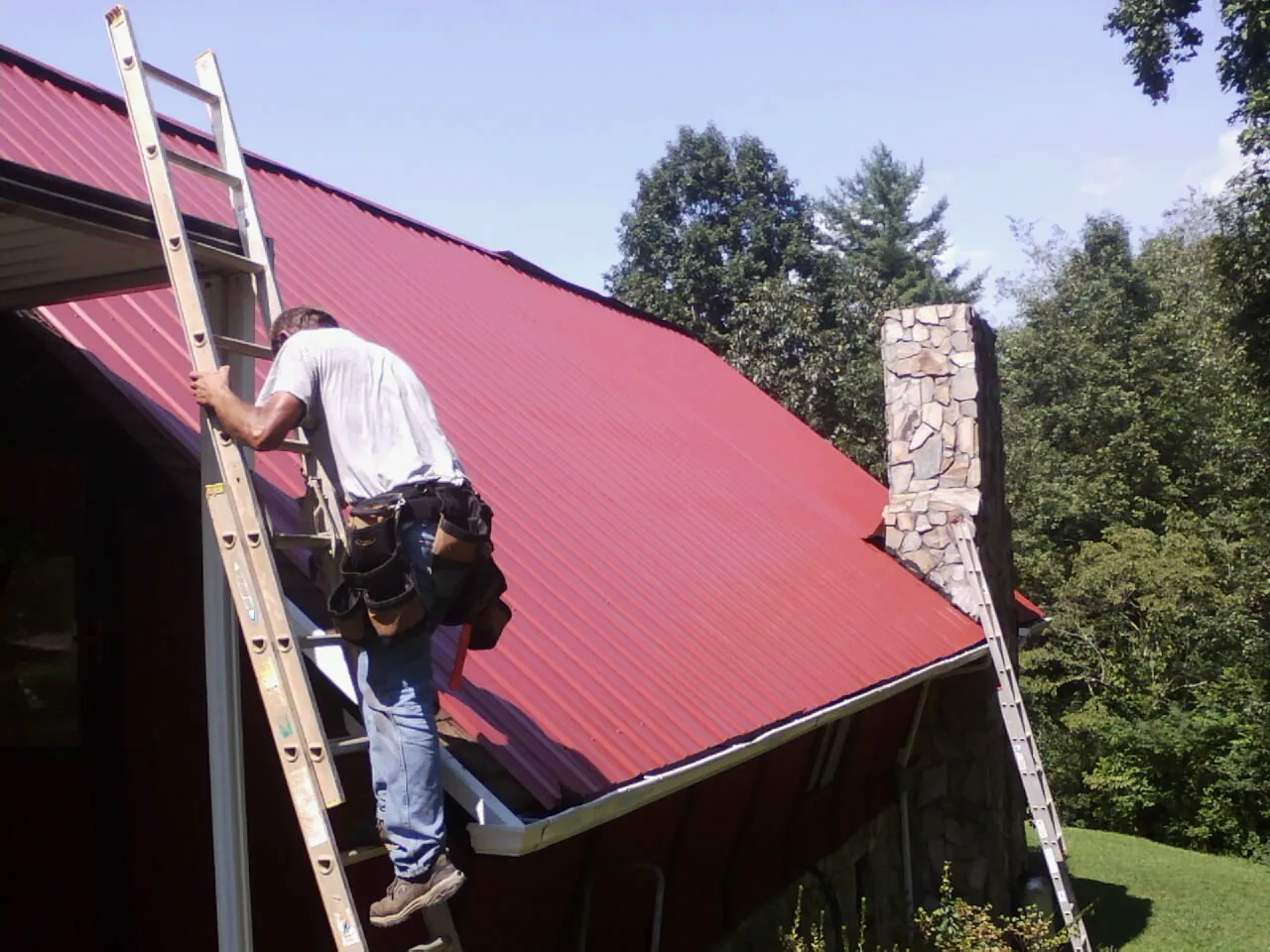 Roofer on a ladder beside a red metal roof and stone chimney — Soffit & Fascia Repair by Roofing Brothers in Lakebay